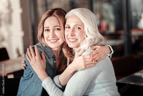 Warm hugs. Emotional young cheerful woman looking glad while being in a nice cafe and hugging her kind beautiful loving grandmother