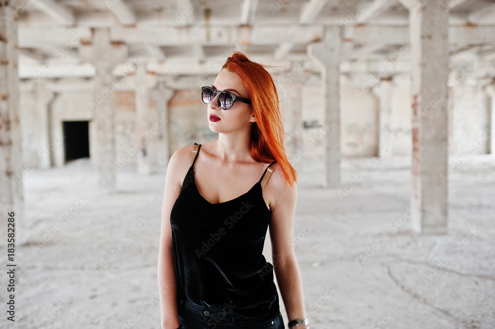 Red haired stylish girl in sunglasses wear in black, against abadoned place.