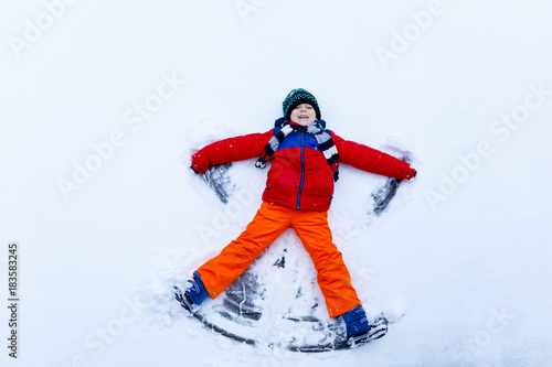 Cute little kid boy in colorful winter clothes making snow angel, laying down on snow