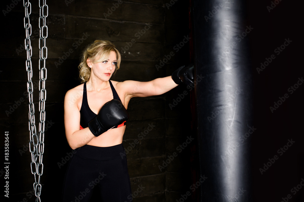 Woman hits the heavy bag with a strong kick. Concept about fit boxe, martial arts and sport. Simple woman in black boxing gloves. She is fighter for success.