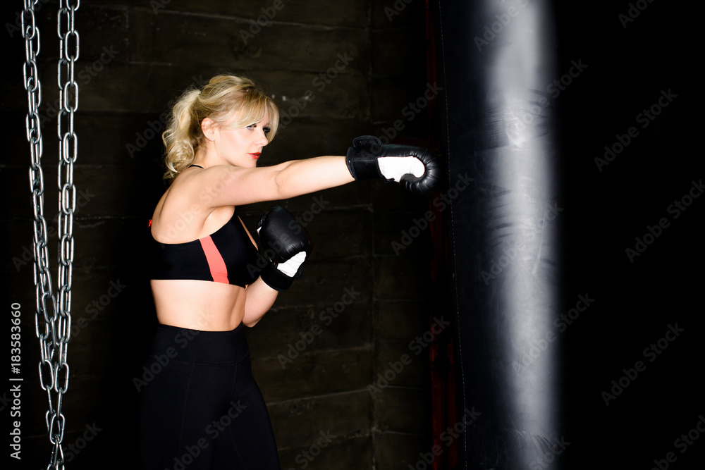 Woman hits the heavy bag with a strong kick. Concept about fit boxe, martial arts and sport. Simple woman in black boxing gloves. She is fighter for success.