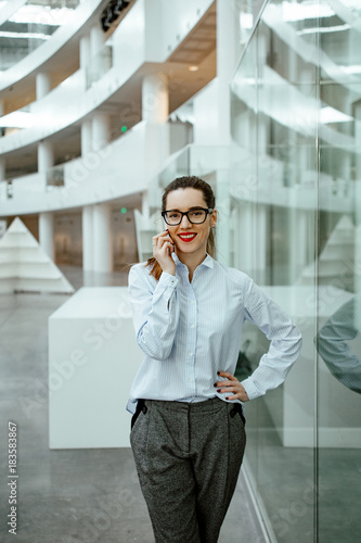 the business lady in strict glasses and a trouser suit talks on the smartphone against the background of business center. the girl smiles and looks in a shot