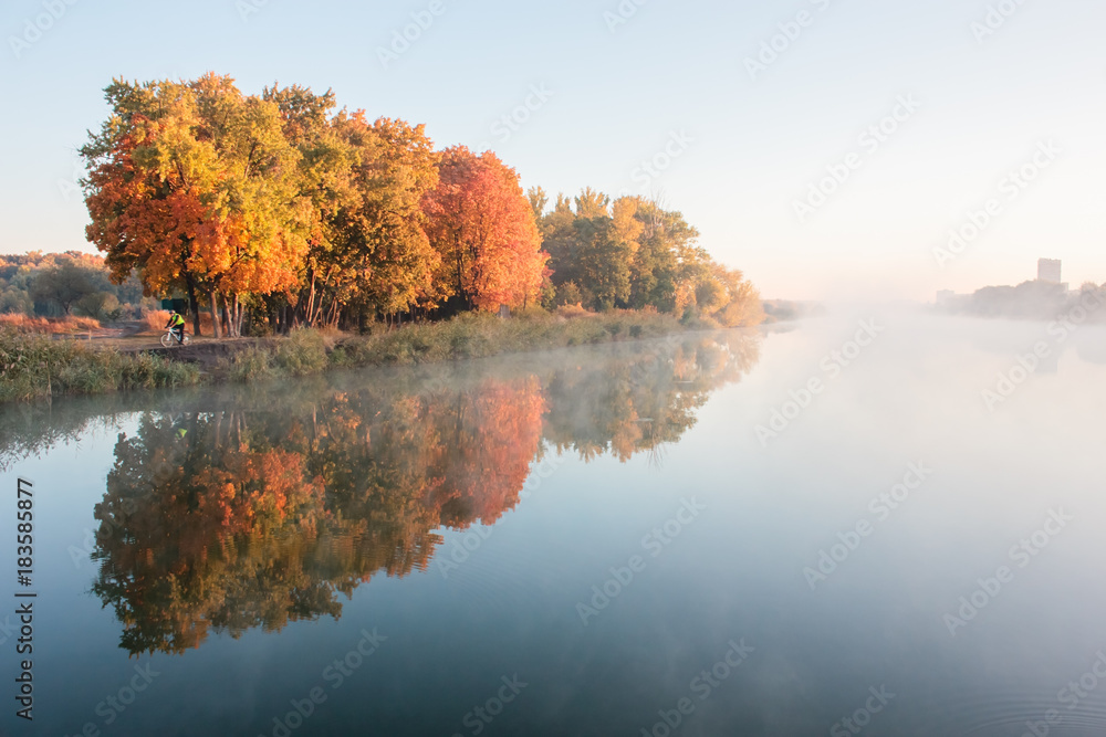 Fototapeta premium Autumn landscape in city park. Heavy fog on river in morning. Orange trees on river bank reflected in water