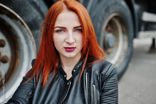 Red haired stylish girl wear in black, sitting against large truck wheels.