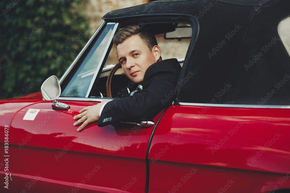 Handsome man in black suit sits in a red retro Italian car