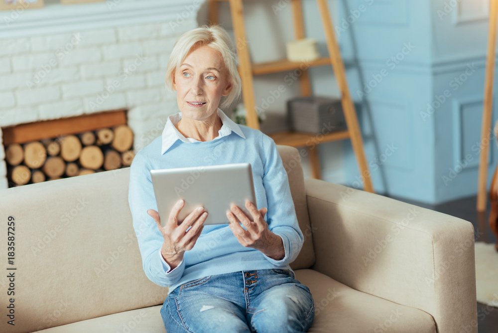 Thoughtful pensioner. Thoughtful senior woman looking into the distance while sitting on a comfortable sofa and holding a wonderful modern tablet in her hands Thoughtful pensioner. Thoughtful senior woman looking into the distance while sitting on a comfortable sofa and holding a wonderful modern tablet in her hands