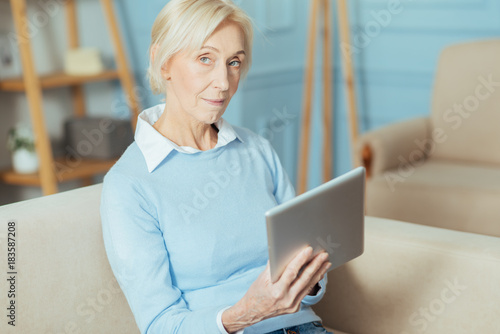 Calm woman. Progressive curious aged woman looking interested while sitting on a sofa and holding a convenient modern tablet in her hands