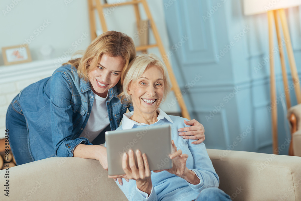 Very funny. Happy senior woman holding a convenient modern tablet and laughing while her granddaughter standing behind her and gently touching her shoulder Very funny. Happy senior woman holding a convenient modern tablet and laughing while her granddaughter standing behind her and gently touching her shoulder