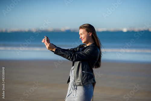 Young woman in a black leather jacket makes selfie on the beach.