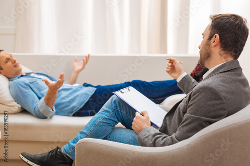 Therapy aids. Experienced professional male psychologist sitting in the armchair and listening to happy male patient who lying on the sofa 
