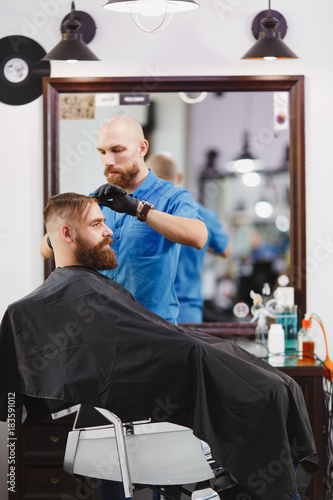 Male professional hairdresser serving client by scissors. Ginger handsome brutal stylish young man with thick big beard, short hair getting trendy haircut in black cape. Light white barber shop room.