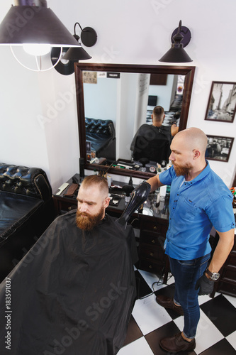 Male professional hairdresser serving client by hair dryer. Ginger handsome brutal stylish young man with thick big beard short hair getting trendy haircut in black cape. Light white barber shop room