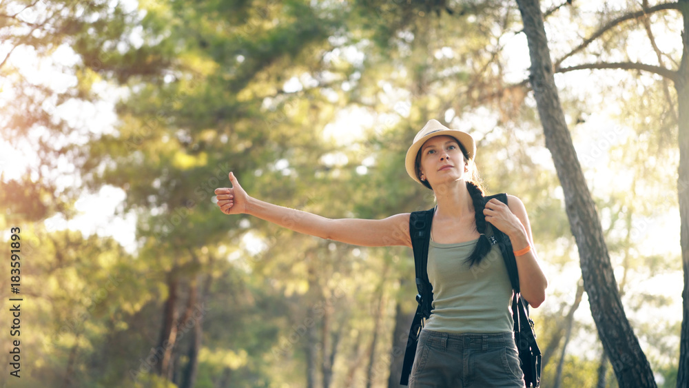Traveler woman hitchhiking on a sunny forest road. Tourist girl looking for ride to start her journey