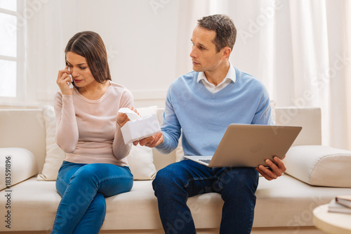 Do not cry. Handsome pleasant man holding laptop and proposing tissue while upset wife crying 