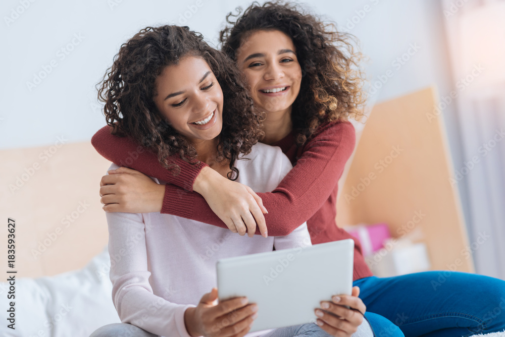 Love her to the moon and back. Extremely happy young lady looking into the camera with a cheerful smile on her face while hugging her sibling looking at a touchpad at home. Love her to the moon and back. Extremely happy young lady looking into the camera with a cheerful smile on her face while hugging her sibling looking at a touchpad at home.