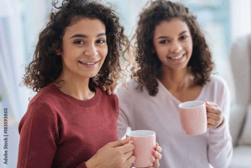 In a positive mood. Selective focus on a beautiful girl in a darker jumper smiling into the camera while sitting next to her best friend and drinking tea at home.