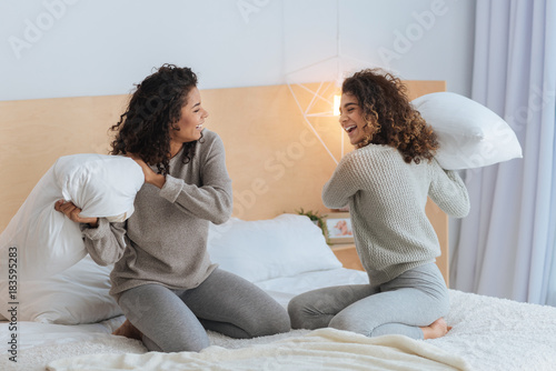 Having so much fun. Excited girls grinning broadly while sitting on a bed and having fun during a family pillow fight at home.