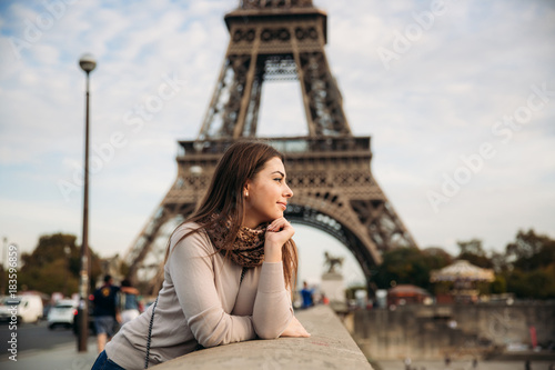 Beautiful girl posing to the photographer against the background of the Eiffel Tower. Autumn photosession. Sunny weather. Beautiful smile and makeup © Aleksandr
