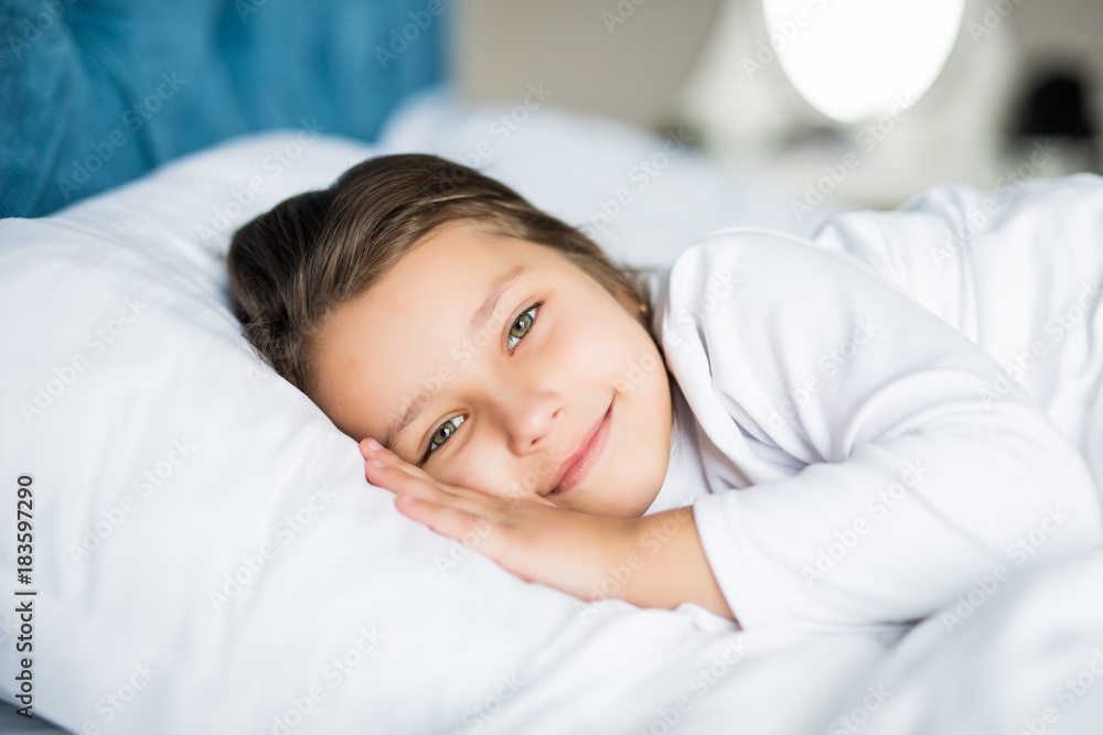 Close-up portrait of laughing little girl lying on bed with hand under pillow, looking at camera and smile. Good morning.