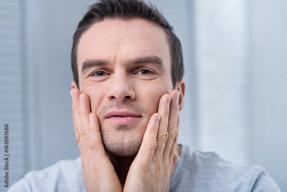 After shaving. Appealing positive pensive man putting lotion after shaving and placing hands on the chicks After shaving. Appealing positive pensive man putting lotion after shaving and placing hands on the chicks