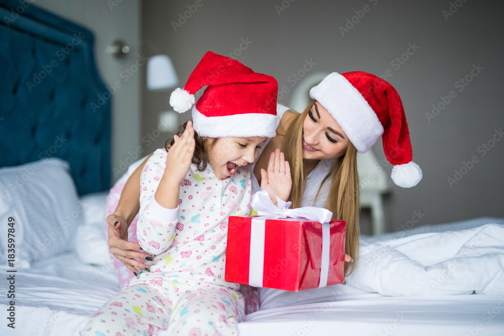 Happy family wearing Santa Claus hats sitting on bed. Mother give and surprised child Santa gift and having fun in Christmas time. Winter holiday Xmas and New Year concept Happy family wearing Santa Claus hats sitting on bed. Mother give and surprised child Santa gift and having fun in Christmas time. Winter holiday Xmas and New Year concept