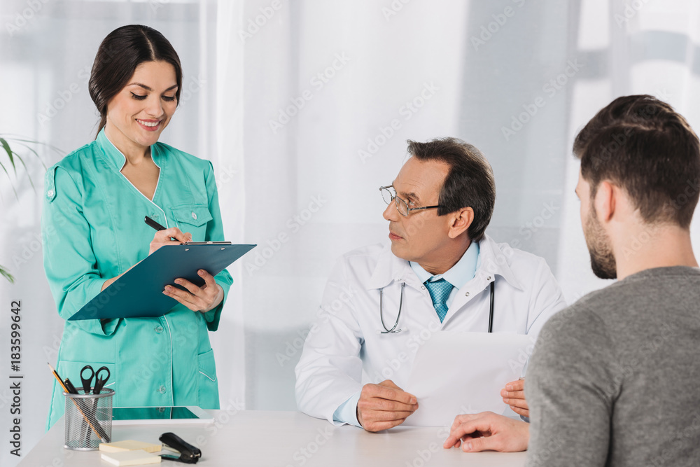 Fototapeta premium doctor looking at smiling nurse writing something to clipboard