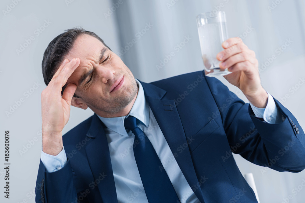 Headache driving crazy. Sad brunette young man closing his eyes while carrying glass of water and touching his forehead Headache driving crazy. Sad brunette young man closing his eyes while carrying glass of water and touching his forehead