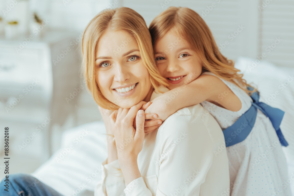 Our family. Cute smiley positive girl standing behind her mother and hugging her while being in a great mood Our family. Cute smiley positive girl standing behind her mother and hugging her while being in a great mood