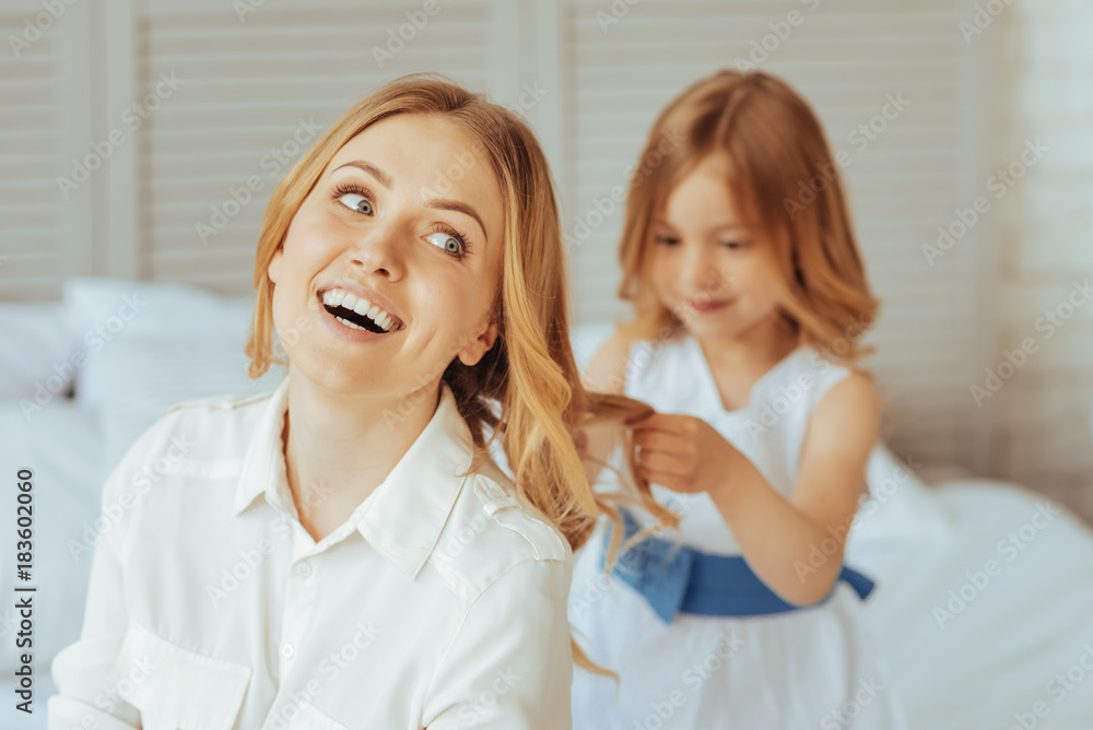 New hairstyle. Delighted positive creative girl sitting behind her mother and holding her hair while doing a new hairstyle for her