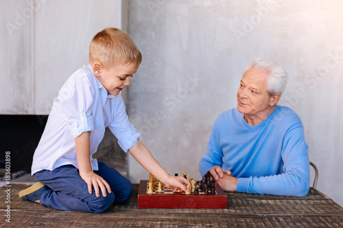 Developing chess. Delighted nice curious boy holding a chess piece and making a move while leaning to play chess with his grandfather