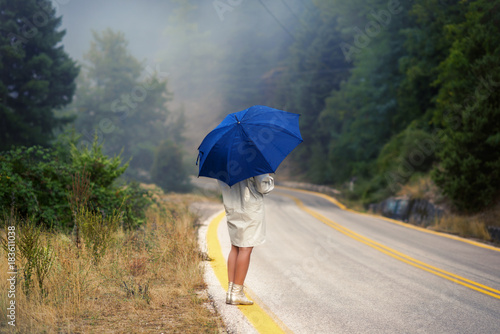 Young female in a raincoat and umbrella on the road in the fog. Travel of women in the raincoat hitchhiking in the rain