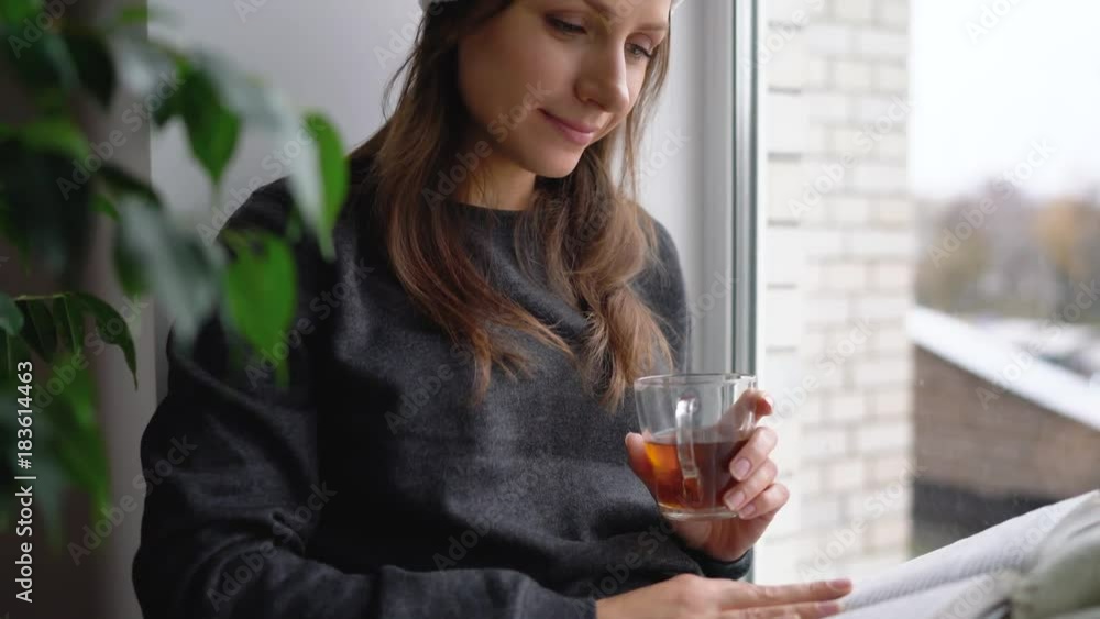 Beautiful young woman in the Santa Claus hat sitting on window sill with tea and reading book in anticipation of Christmas Beautiful young woman in the Santa Claus hat sitting on window sill with tea and reading book in anticipation of Christmas