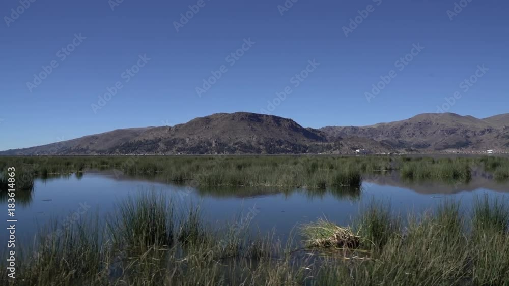 Driving through Uros Islands of high elevation Lake Titicaca in Peru. Calm morning, blue sky, mountain in background. Driving through Uros Islands of high elevation Lake Titicaca in Peru. Calm morning, blue sky, mountain in background.