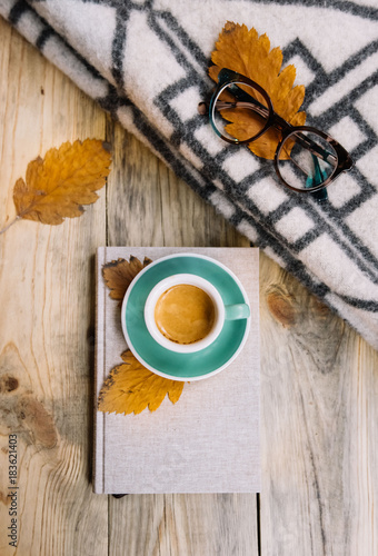 Autumn setup: a green ceramic cup of fresh morning espresso coffee with a tiger crema standing on a book; reading glasses and warm wool blanket on the rustic wooden table background 