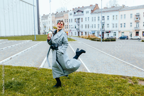 Young fashionable woman blogger in gray cashmere coat in sunglasses, coffee cup in hand, enjoying and jumping over ciyu view background. photo