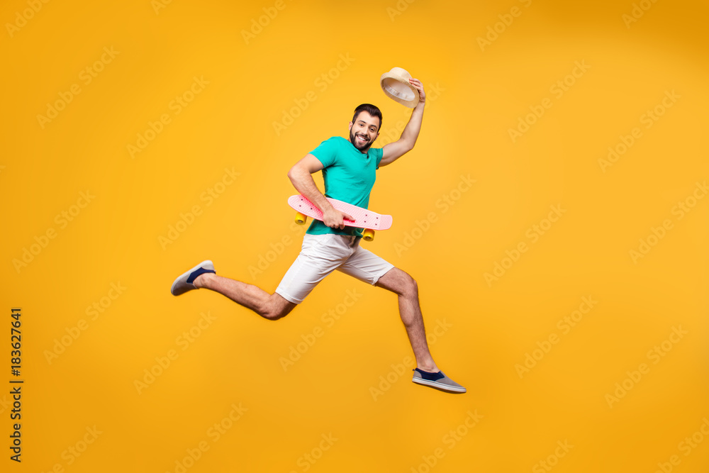 Funny smiling happy cheerful glad guy is sending his greetings, raising up his hat, he is jumping up with skateboard in hands, isolated on yellow background, copyspace