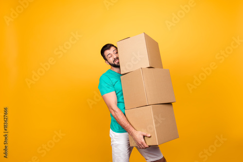 Concept of courier and messenger service. Grimacing handsome guy is trying to keep big stack of cardboard bosex in his hand, isolated on yellow background