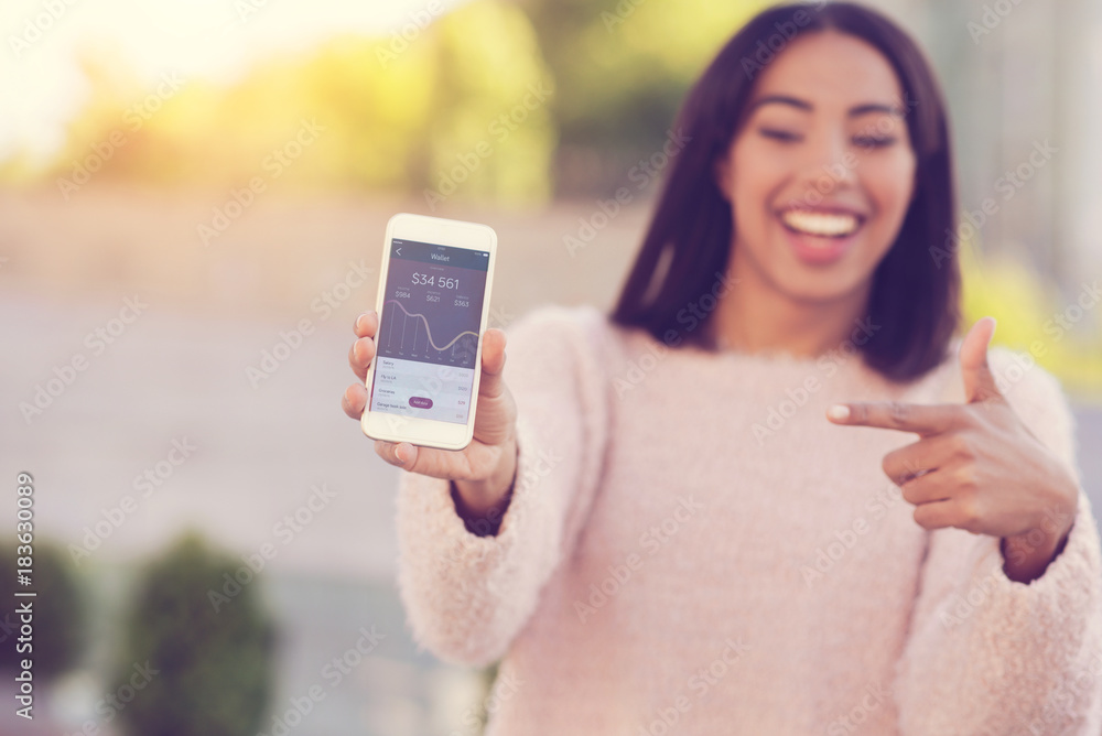 Modern app. Selective focus of a modern smartphone being in hands of a pleasant delighted young woman while pointing at it