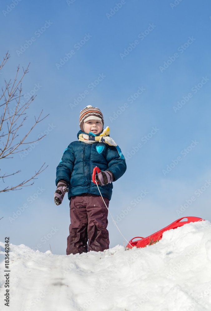 Boy standing with red plastic sledge on a snowy hill Boy standing with red plastic sledge on a snowy hill
