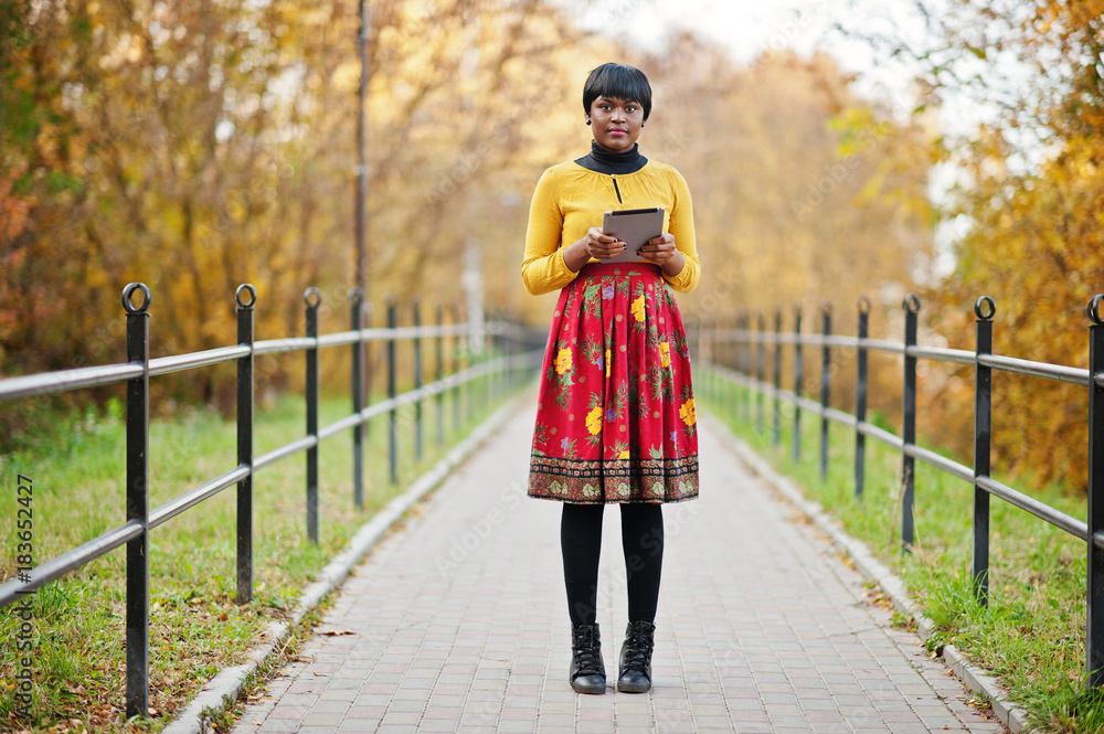 African american girl at yellow and red dress at golden autumn fall park with tablet at hands.