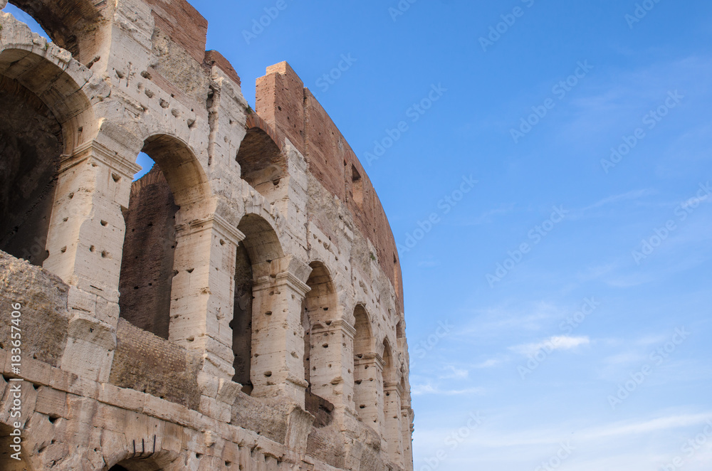 A corner of the top level of the Colosseum in Rome against a blue sky with white clouds floating lazily above