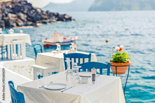Fototapeta Grecja, Santorini. Restauracja ze stolikiem serwowanym w nadmorskim wybrzeżu Morza Egejskiego na wyspie Cyclades Santorini z zapierającym dech w piersiach, niesamowitym i niewiarygodnym widokiem na wodę i nabrzeże wioski Oia Ia.