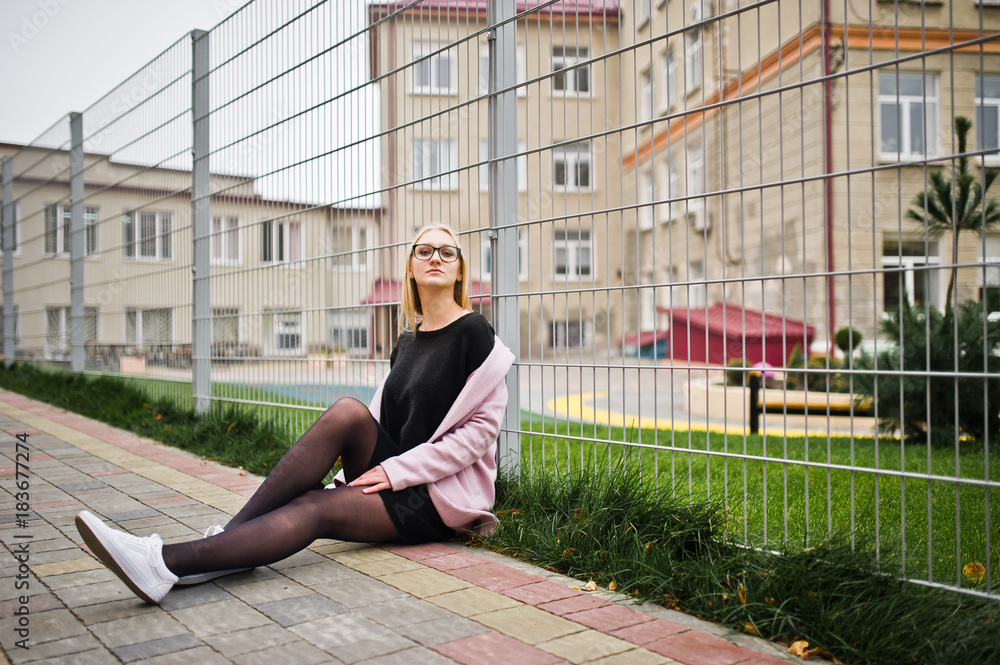 Blonde girl at glasses and pink coat, black tunic sitting against fence at street.
