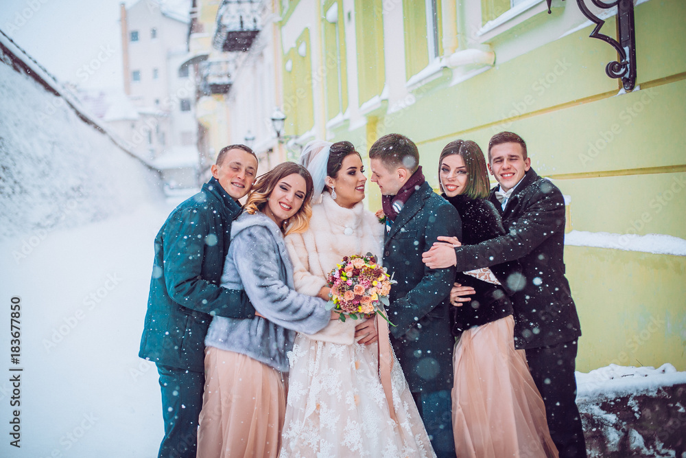 Bridesmaids and groomsmen dressed in powder dress tones surround stylish wedding couple in winter snowy street.