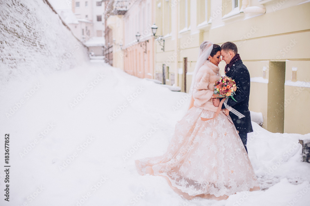 Beautiful bride and groom standing on a snowy street. Newlyweds kiss.