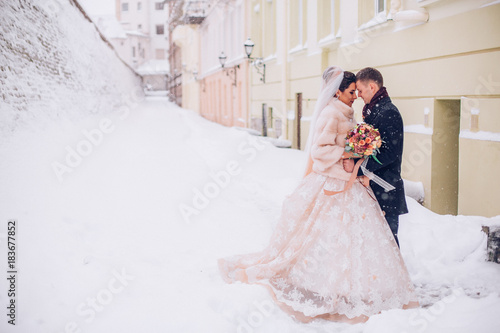 Obraz na płótnie Beautiful bride and groom standing on a snowy street. Newlyweds kiss. w salonie