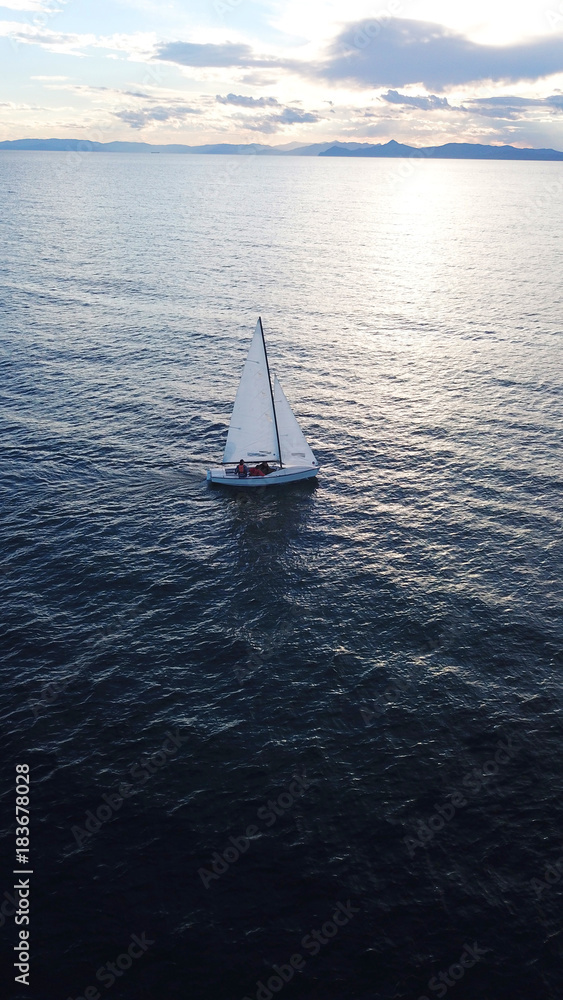 Fototapeta premium Aerial drone bird's eye view of yacht cruising near small islet of Ydrousa at sunset with beautiful scattered clouds, Voula, Athens riviera, Attica, Greece