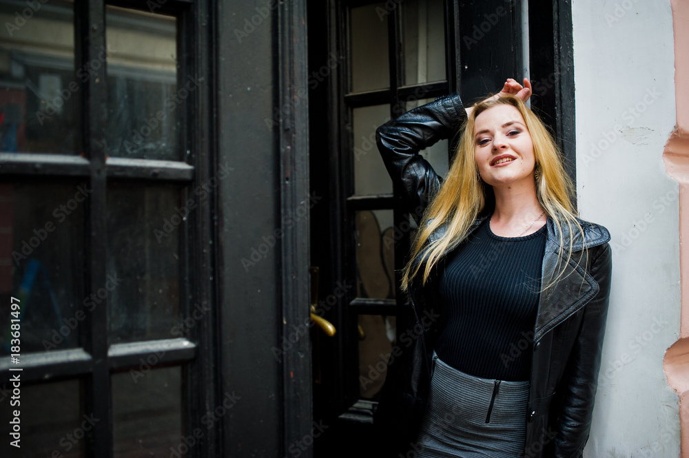 Fototapeta premium Blonde fashionable girl in long black leather coat posed against wooden door at old house.