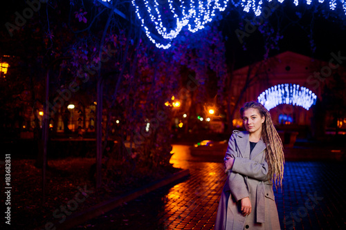 Girl with dreadlocks walking at night street of city against garland lights.