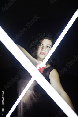 Curly brunette plus size model with two long led lamp at black room.